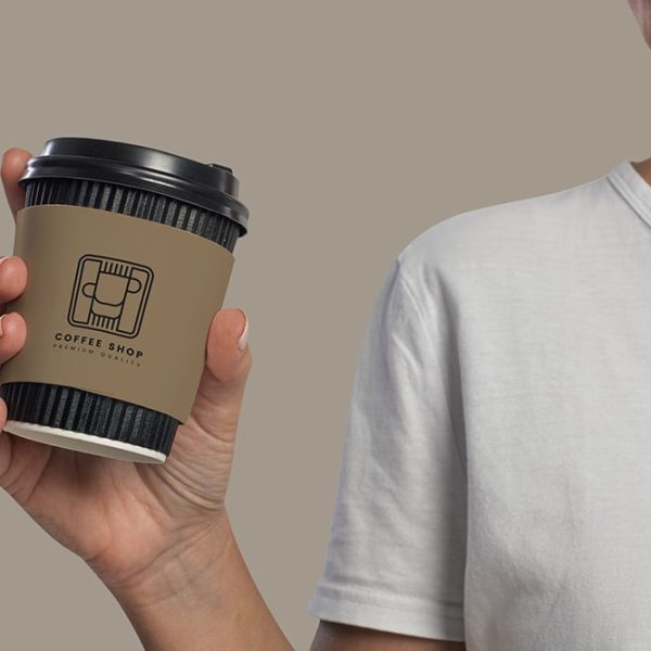 A person wearing a white t-shirt holds a coffee cup with a brown sleeve featuring the logo of "Coffee Shop." The cup has a black lid, and the background is a neutral beige color. The scene seems ideal for sharing on your favorite mobile app. The person's face is partially visible, turned to the right.