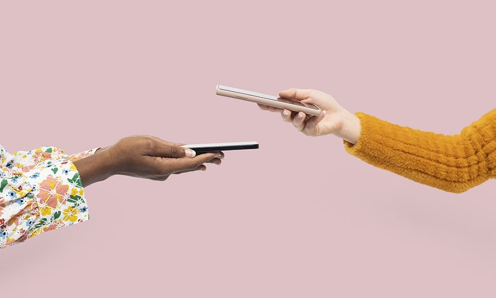 Two hands exchanging smartphones against a pink background. The left hand, with a vibrant floral sleeve, holds a black phone, while the right hand, in an orange sweater, offers a silver phone. The moment of Mobile Sharing is captured mid-air.